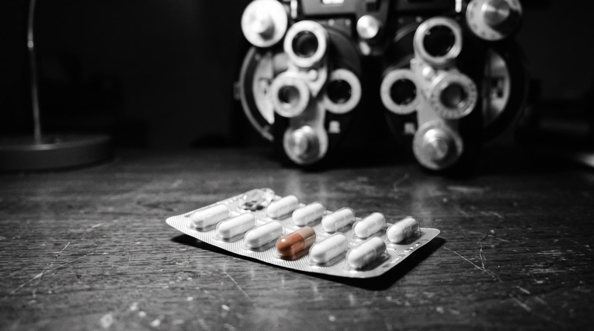 A blister pack of capsules with a blurred eye-exam instrument behind (photo illustration)